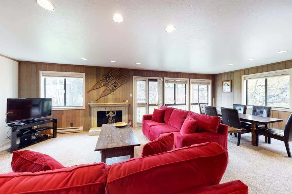 Sala de estar con asientos rojos, un televisor y una mesa de comedor junto a las ventanas.