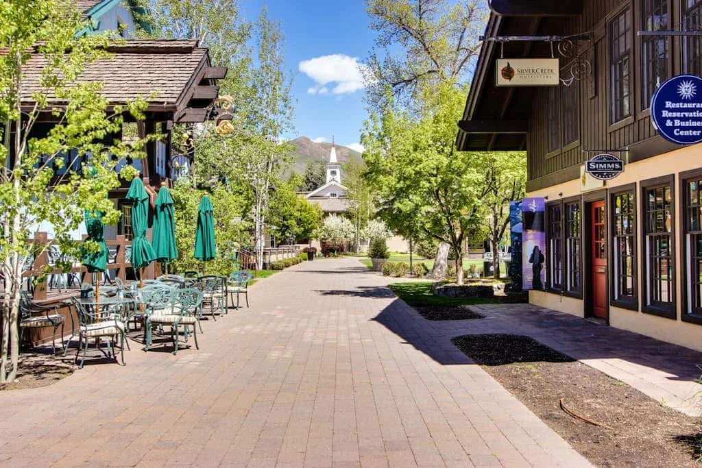 Una calle peatonal pavimentada pasa entre tiendas, mesas al aire libre y árboles frondosos.