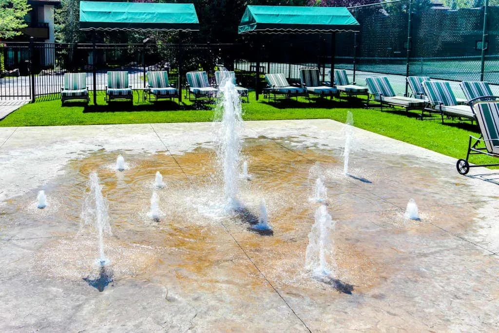 Una zona de juegos acuáticos con chorros de agua está junto a un césped con tumbonas.