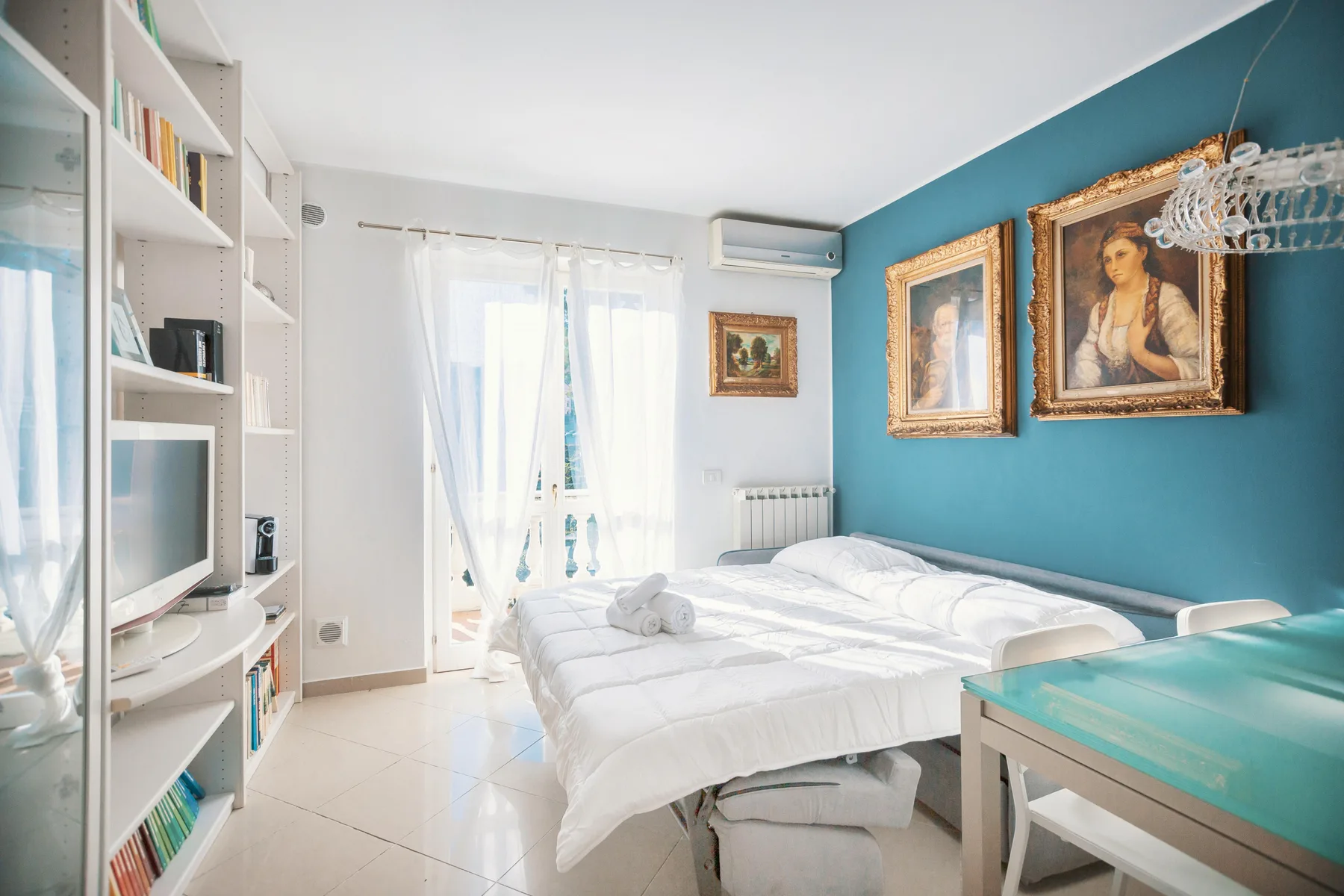 A bright bedroom with white bedding, a turquoise accent wall, and framed artwork.