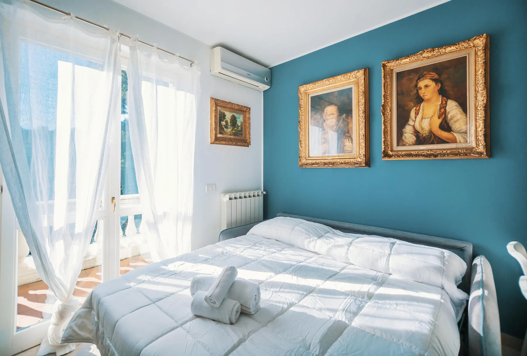 A bedroom with a bed, sheer curtains, and framed artwork on a blue wall.