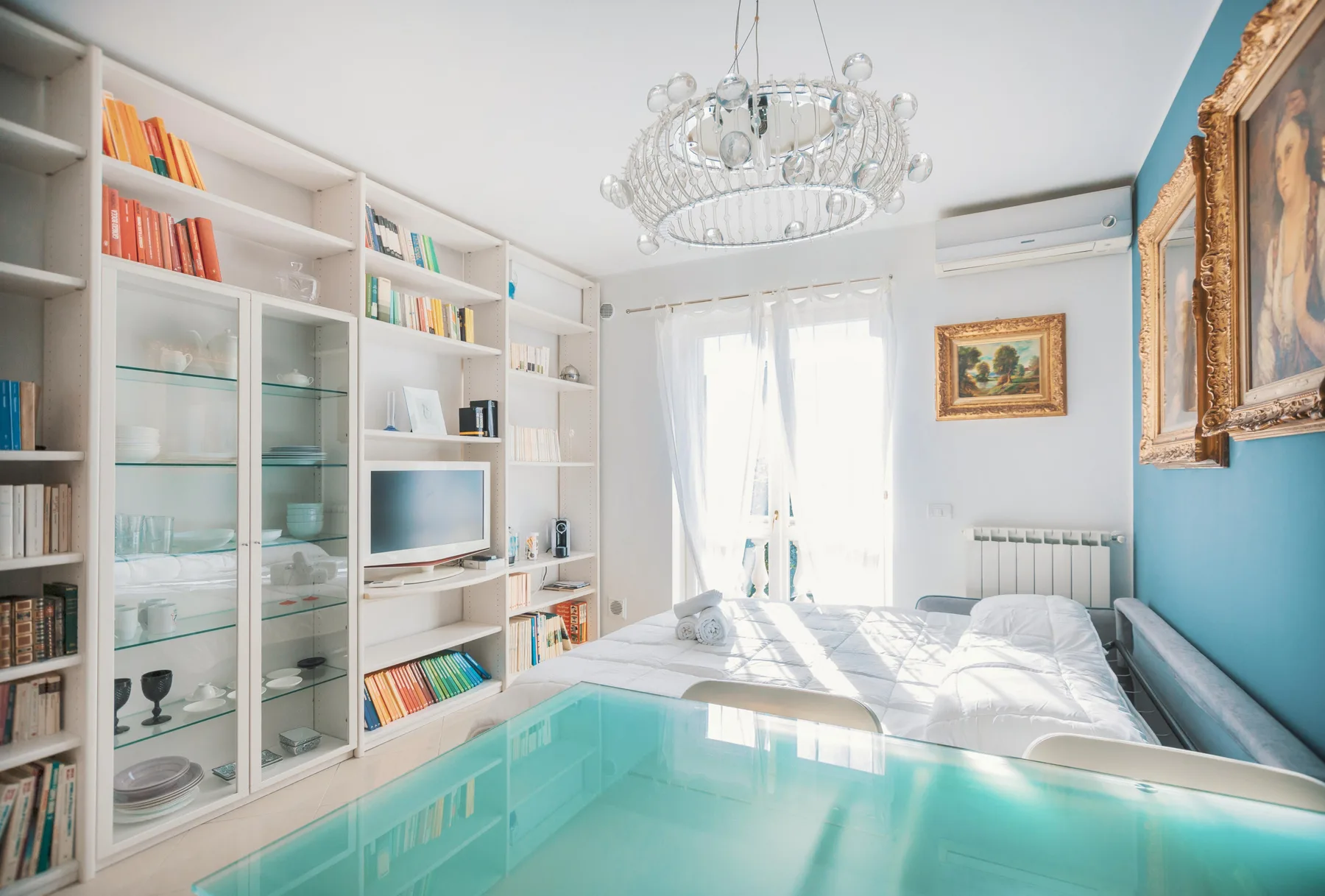 A bright bedroom with built-in shelving, a bed, and a glass chandelier.