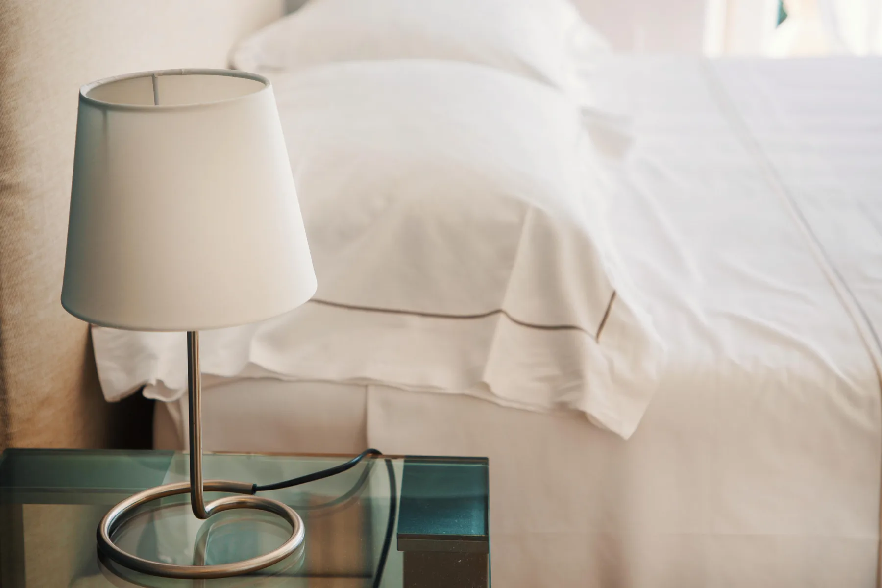 A bedroom with a white bed and a lamp on a glass bedside table.