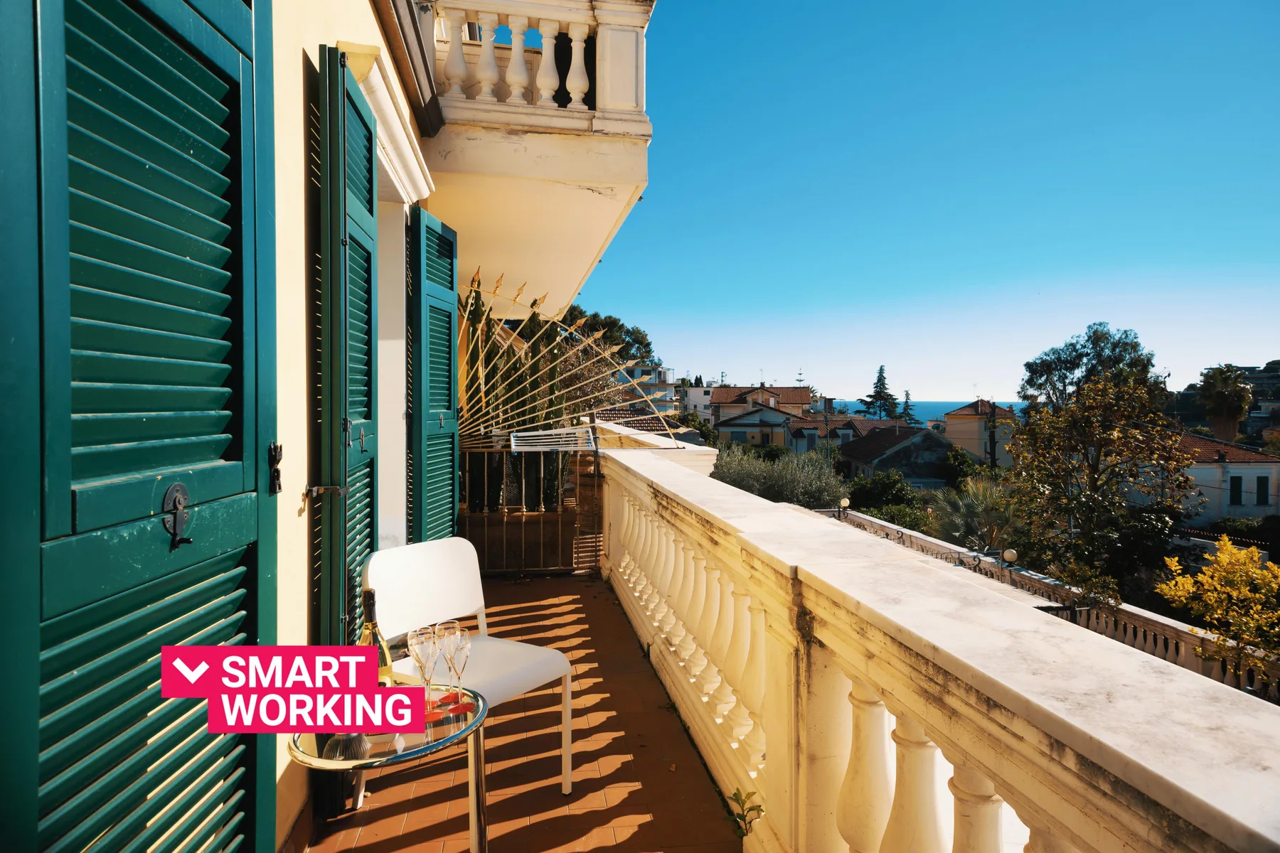 A sunlit balcony with green shutters, a railing, and a view of nearby rooftops.