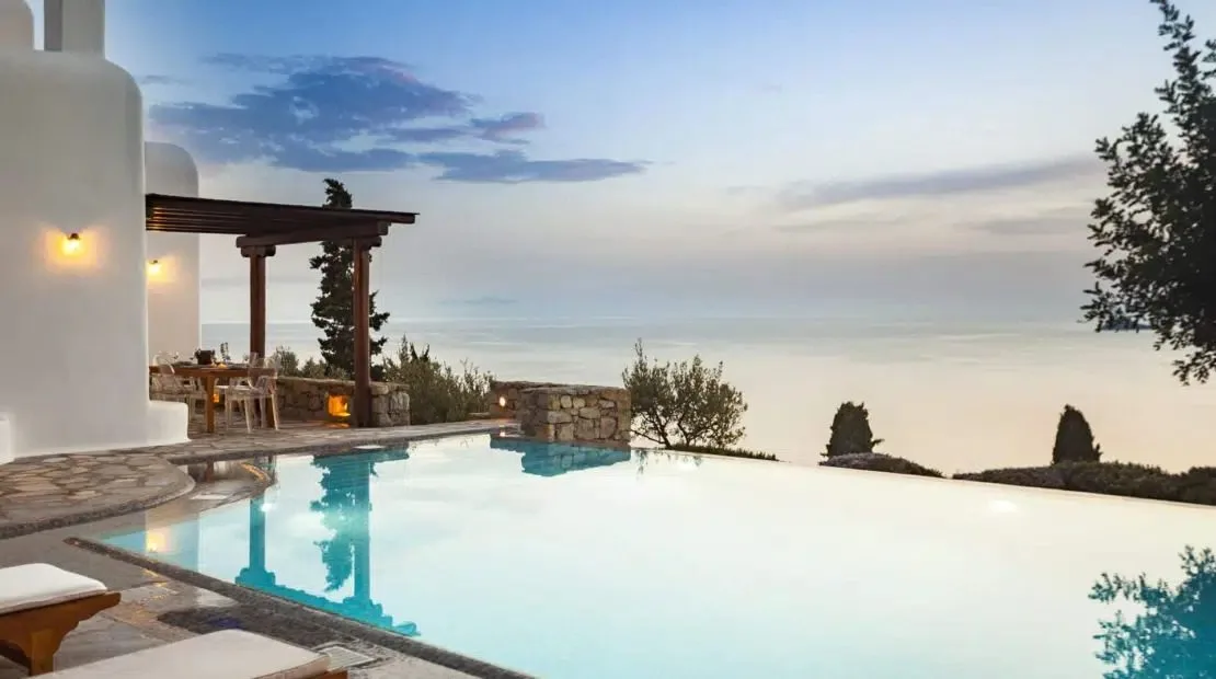 An infinity pool overlooks a hazy sea beside a shaded outdoor seating area.