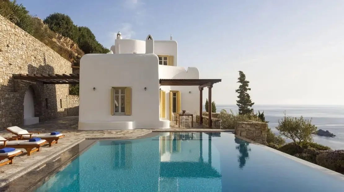 Una villa blanca se alza junto a una piscina, con muros de piedra y vegetación en la ladera.