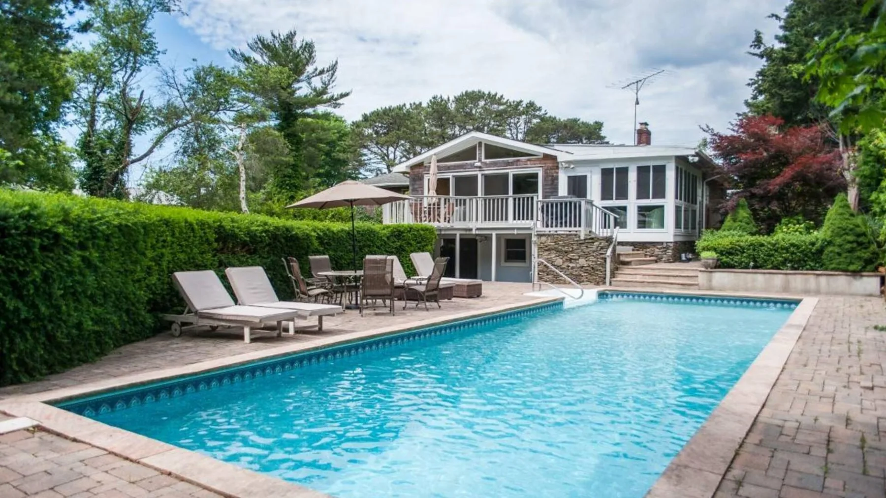 A swimming pool and paved patio sit beside a house and garden.