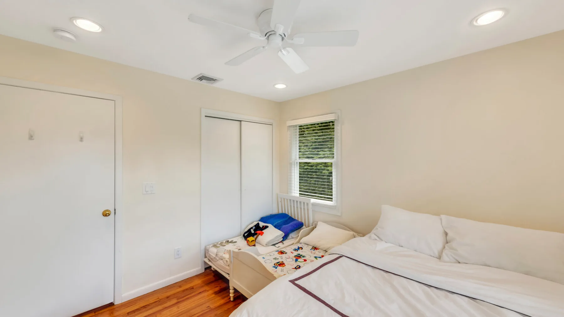 Un dormitorio luminoso con camas, una ventana y un ventilador de techo.