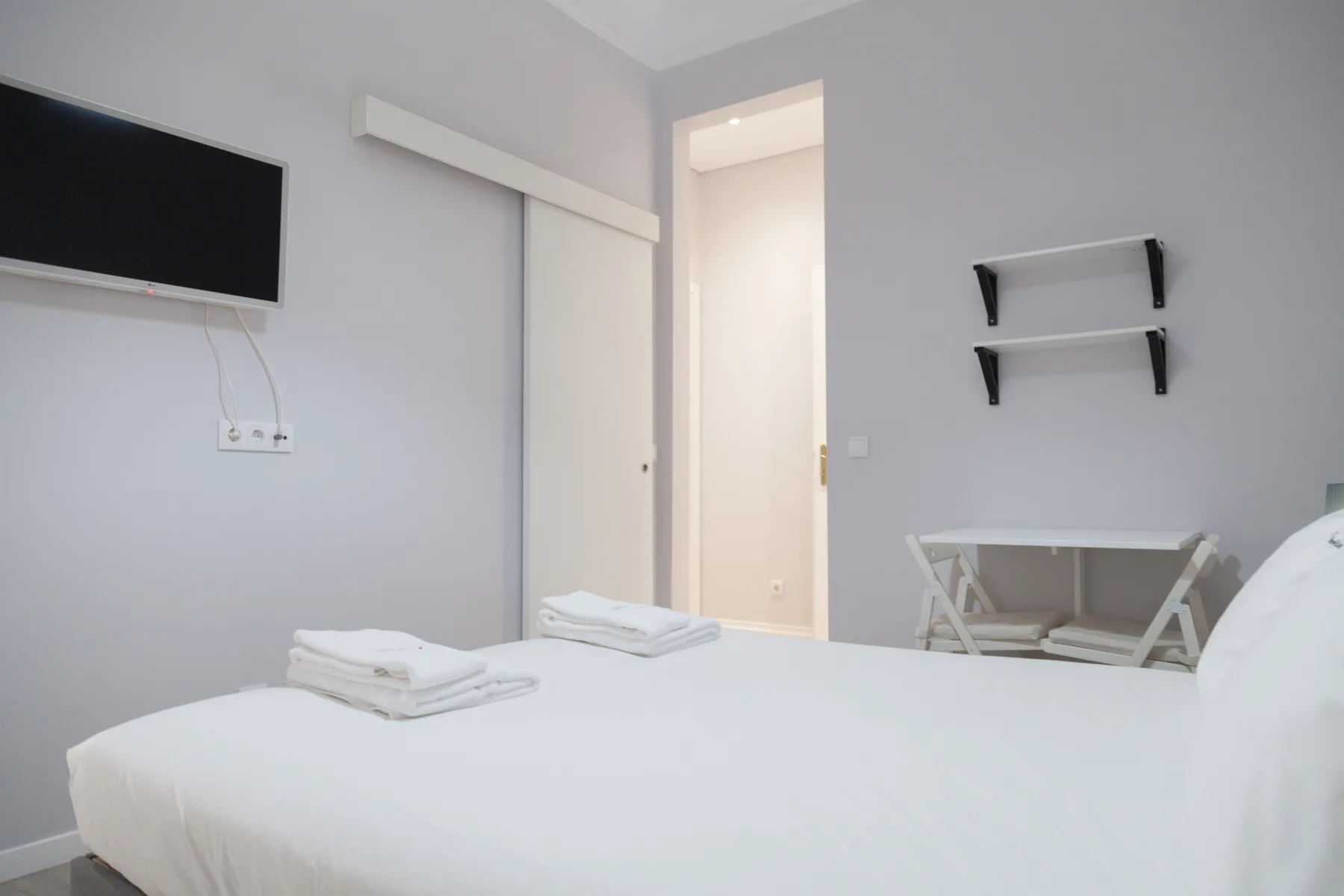Un dormitorio luminoso con una cama bien tendida y un televisor en la pared.