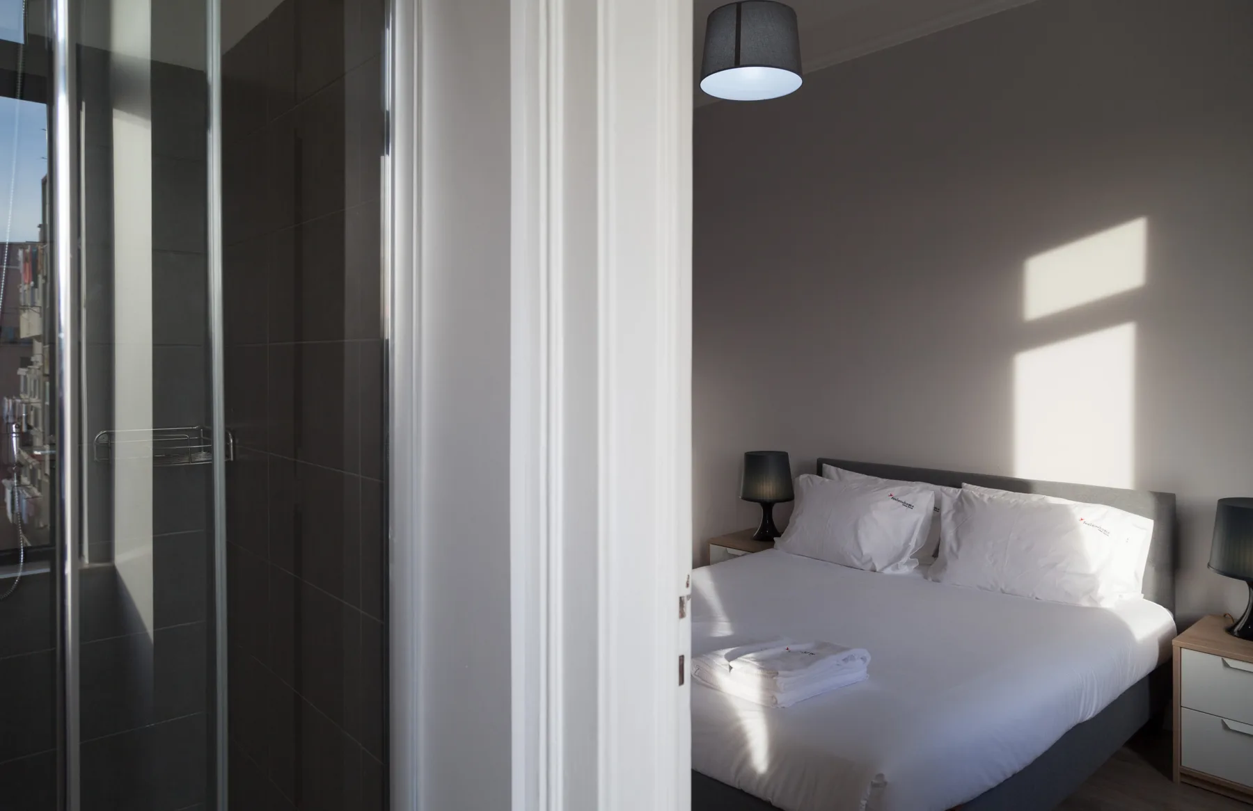 Un dormitorio con una cama bien tendida junto a una ducha de azulejos oscuros.
