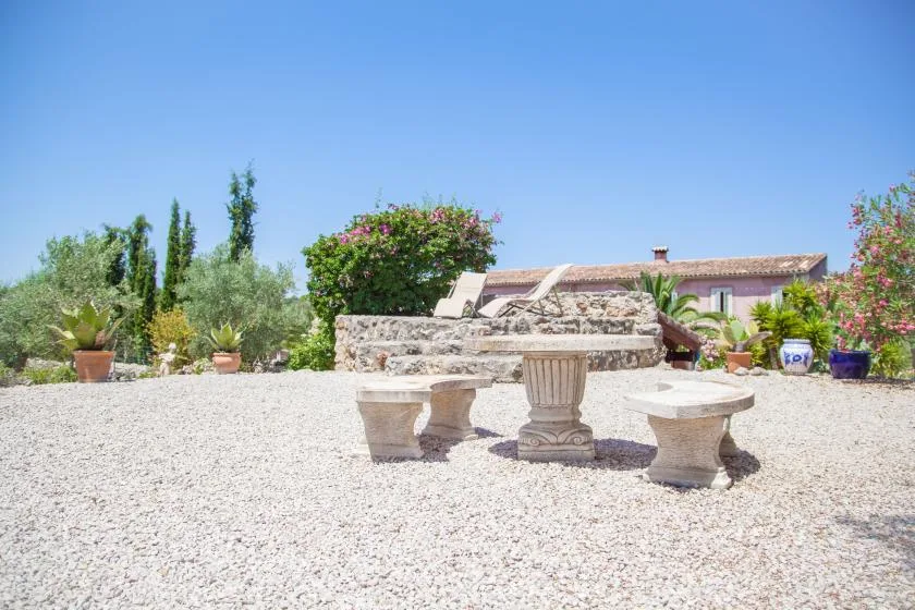 Un patio de grava tiene una mesa de piedra y asientos a juego entre plantas con flores.