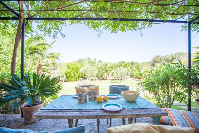 Una mesa de comedor al aire libre, a la sombra, está preparada con vajilla junto a un jardín frondoso.