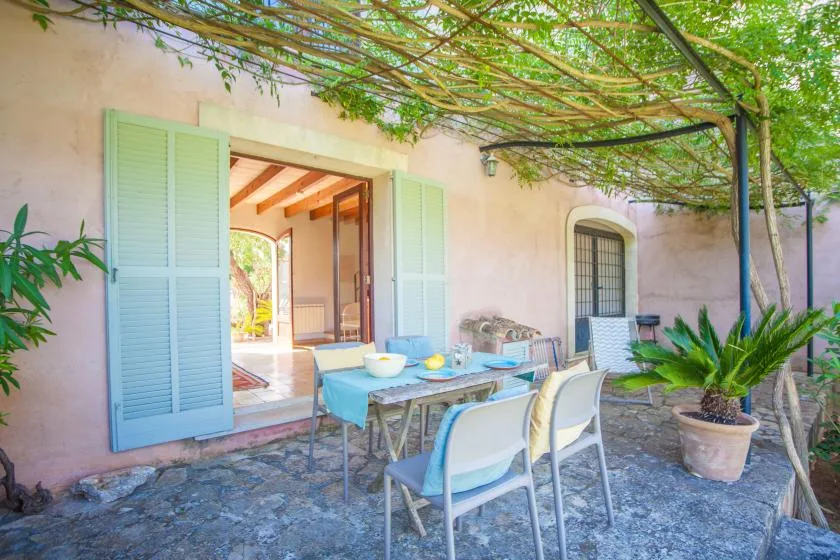 Una zona de comedor al aire libre está junto a puertas abiertas bajo una pérgola frondosa.