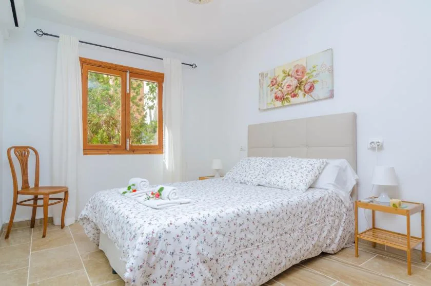 A bedroom with a bed beneath floral wall art and beside a wood-framed window.