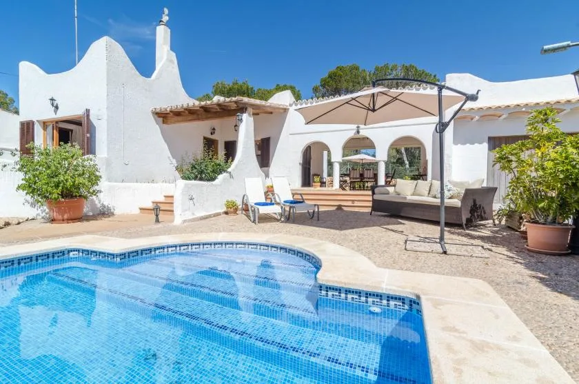 A swimming pool sits beside a white villa patio with outdoor seating and an umbrella.