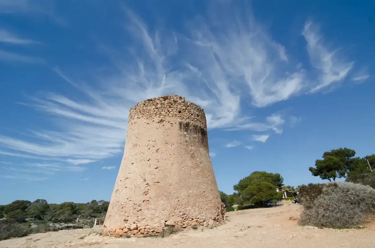 A historic stone tower stands among trees beneath a bright blue sky.