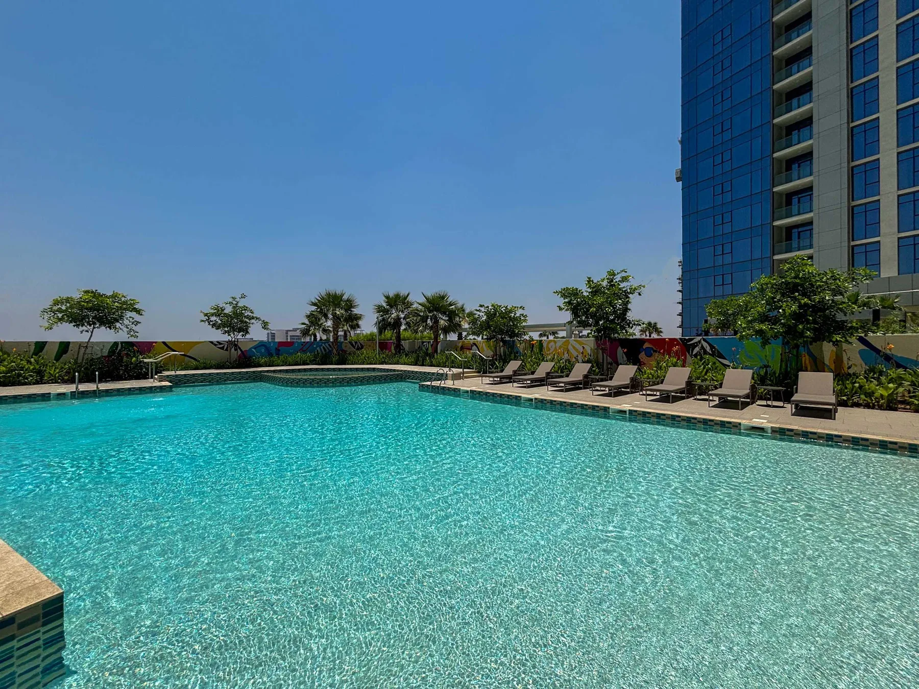 An outdoor swimming pool beside a high-rise building, with lounge seating along the deck.