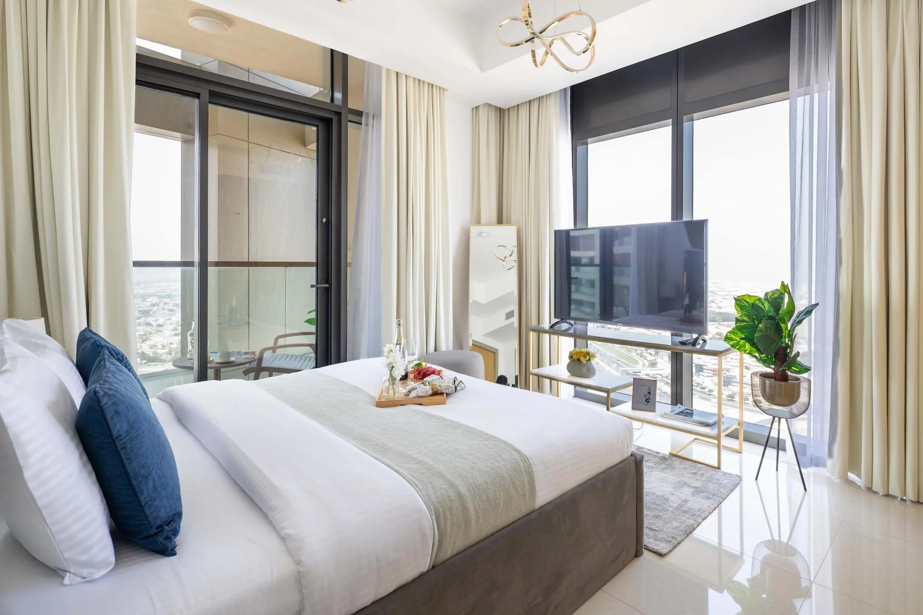 A bright bedroom with a neatly made bed and a television beside large windows.
