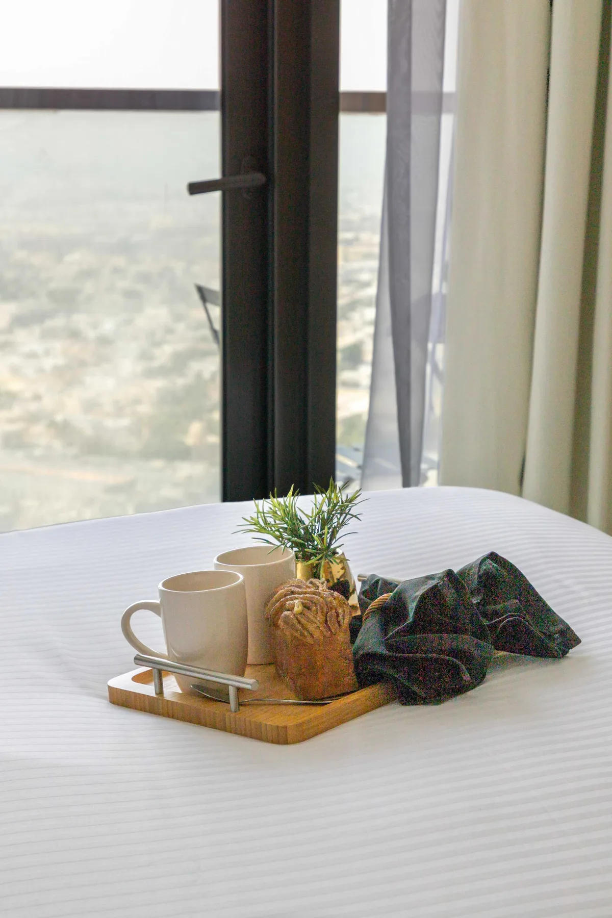A bed by a large window holds a tray with cups, a plant, and folded fabric.