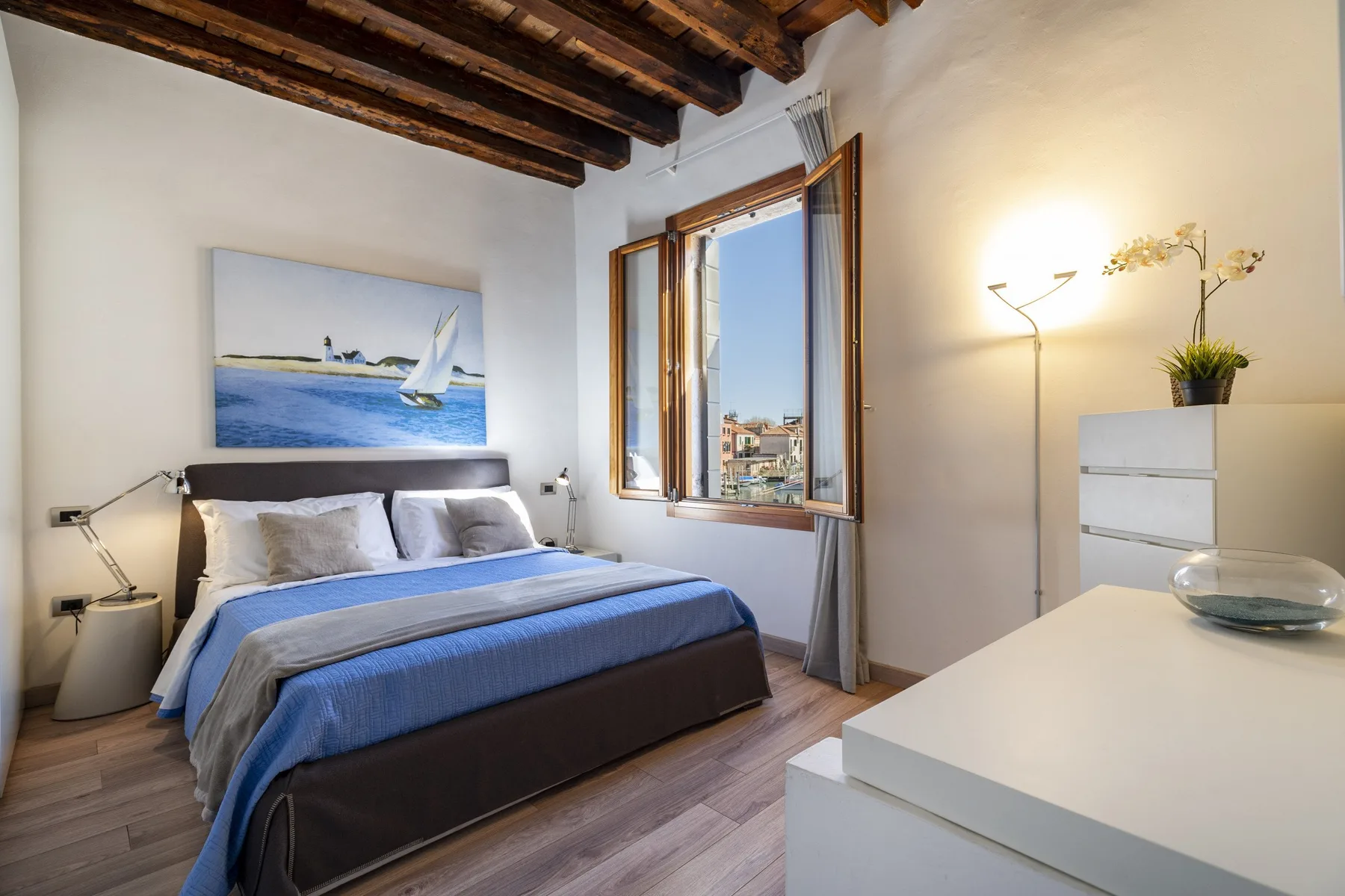 Un dormitorio con una cama bajo un cuadro de un velero y una ventana abierta.