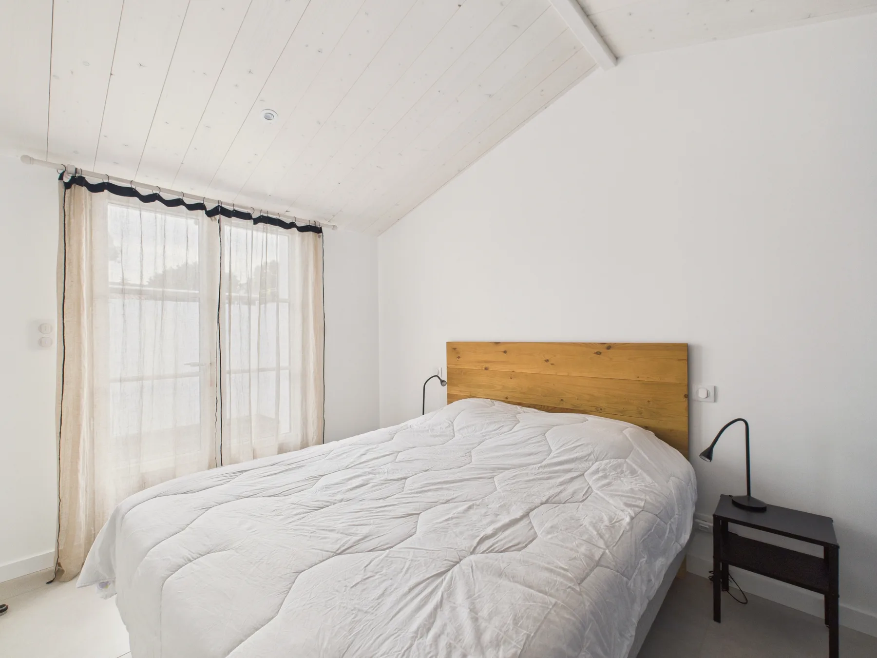 A bright bedroom with a bed beside a curtained window.