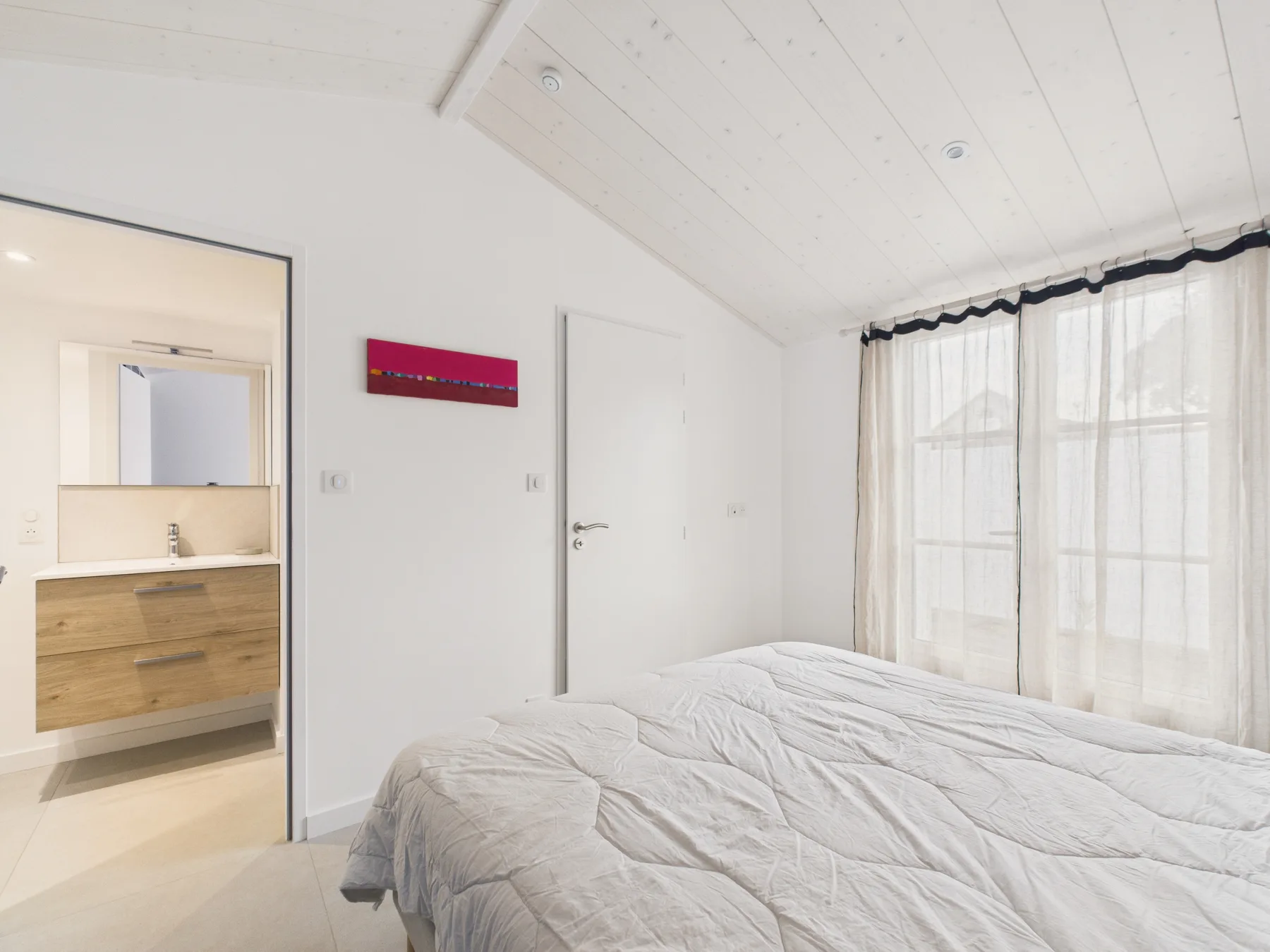 A bright bedroom with a bed beside a curtained window.