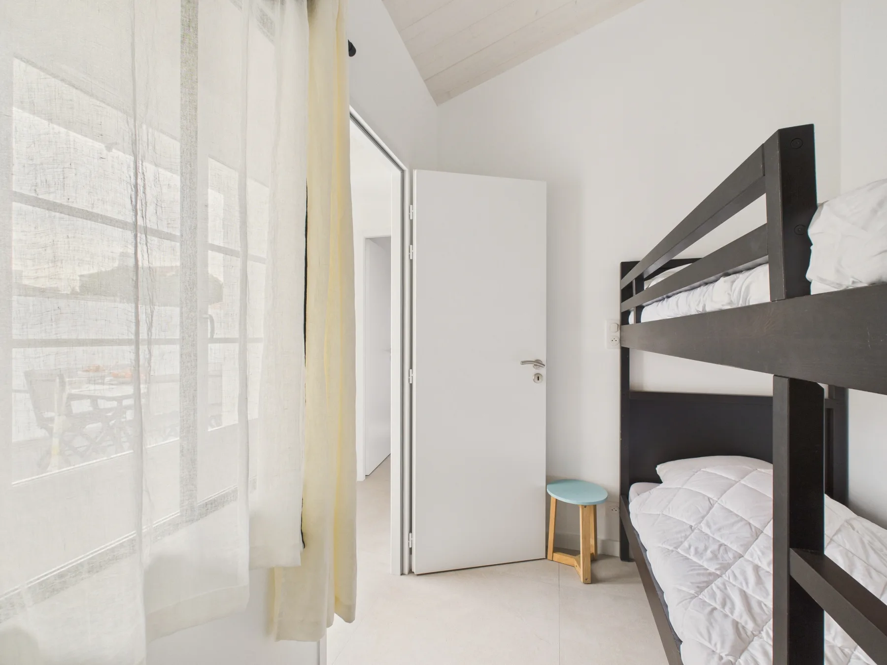 A bright bedroom with a dark bunk bed beside a curtained window.