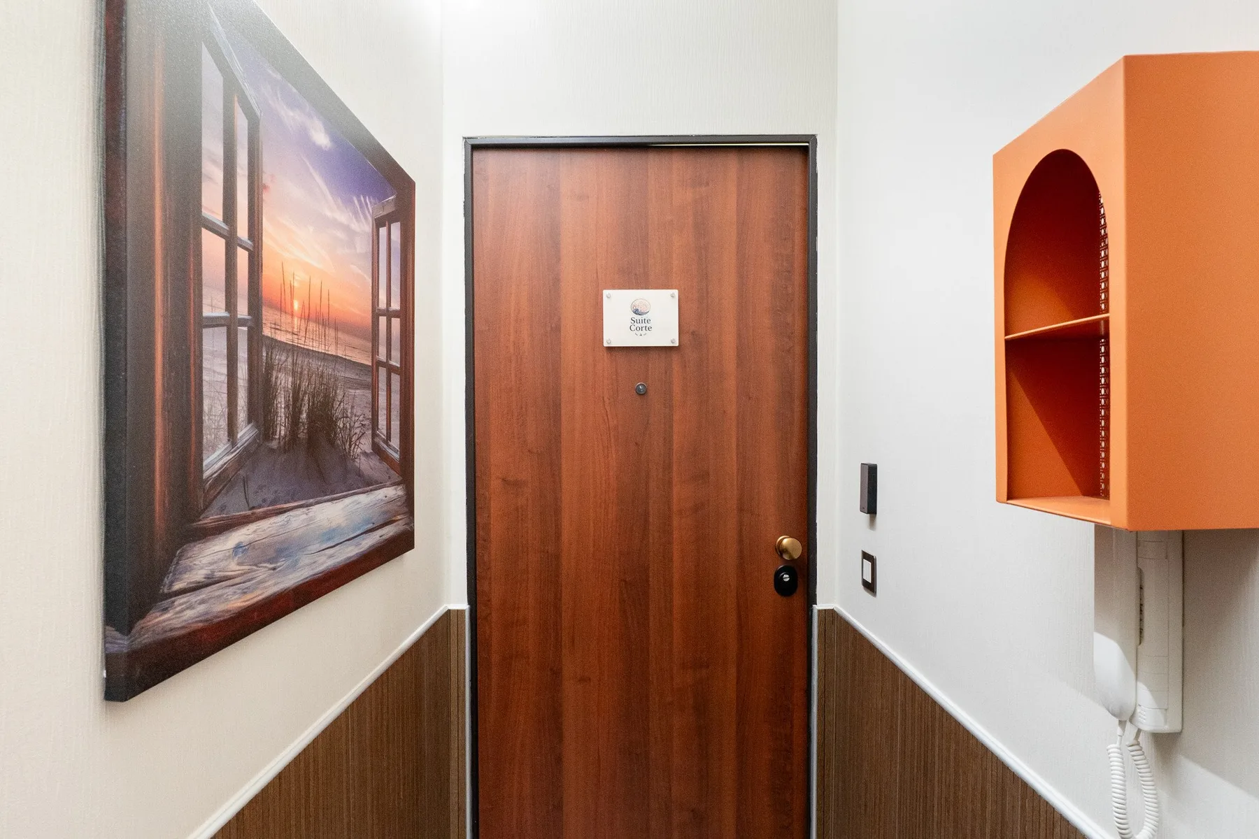 Una entrada con una puerta de madera, una obra enmarcada y un nicho naranja.