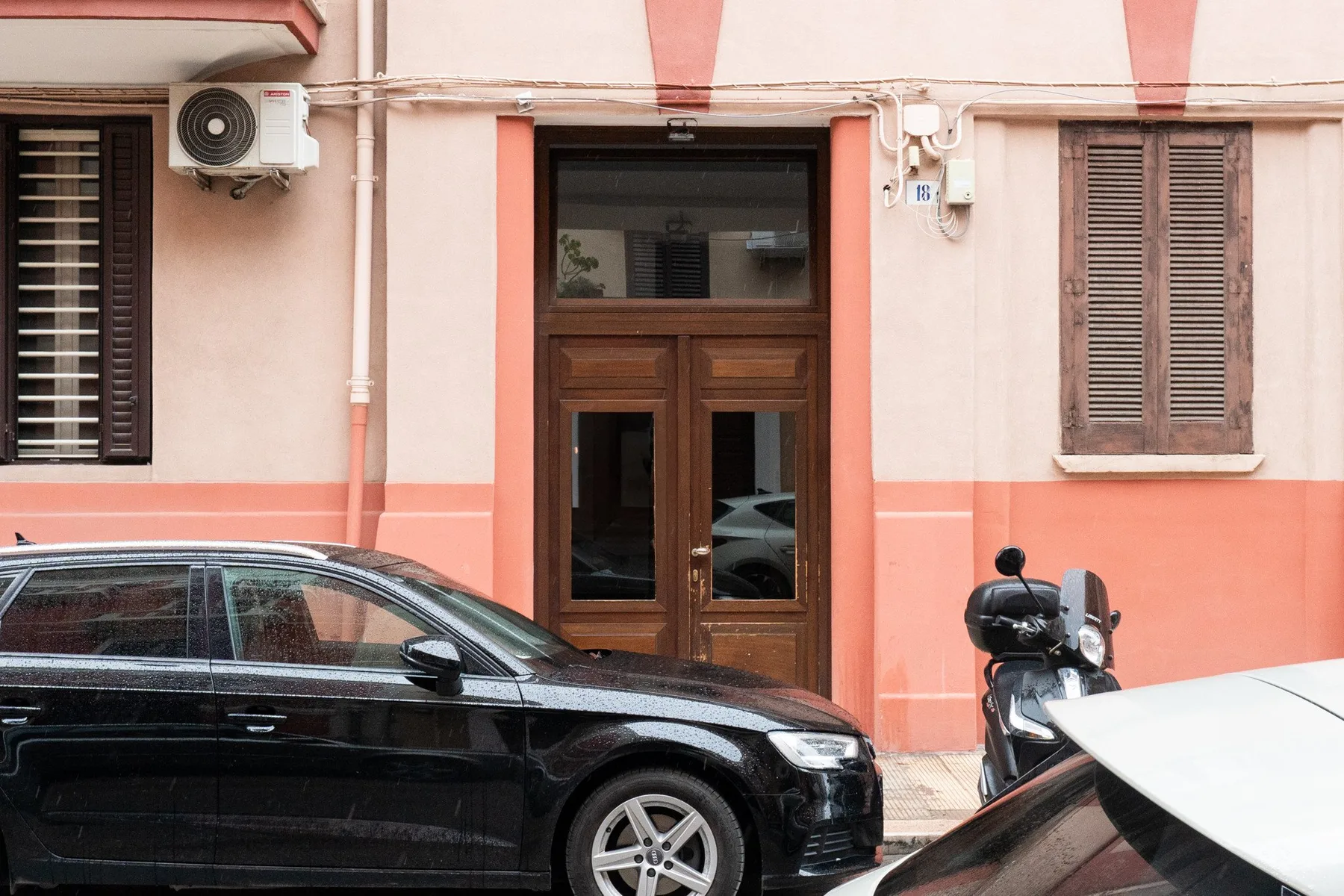 Entrada de un edificio rosa con puerta de madera, coches estacionados y una motoneta.