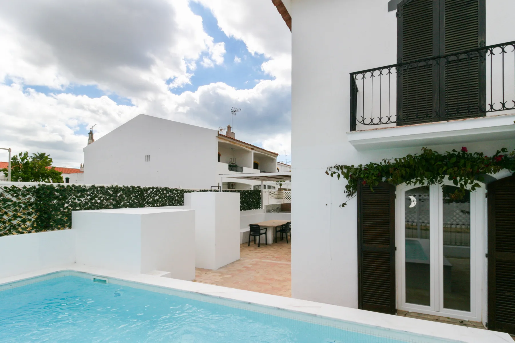 Exterior de una casa junto a una piscina, con contraventanas oscuras y asientos en el patio.