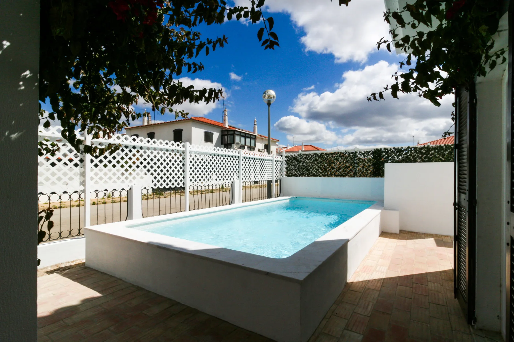Una piscina al aire libre junto a una valla blanca de celosía y un patio pavimentado.