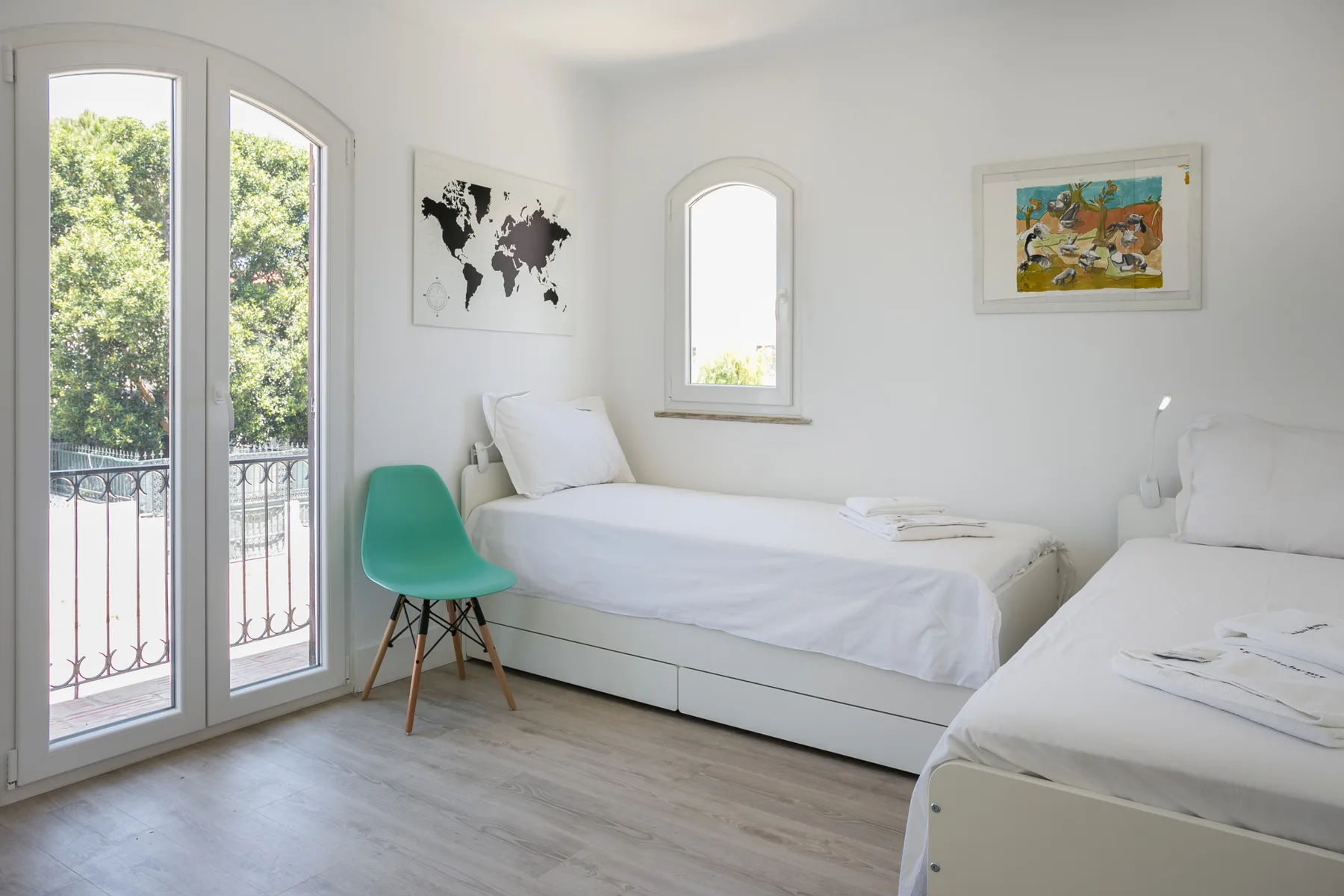 Un dormitorio luminoso tiene dos camas, una silla verde y ventanas altas.
