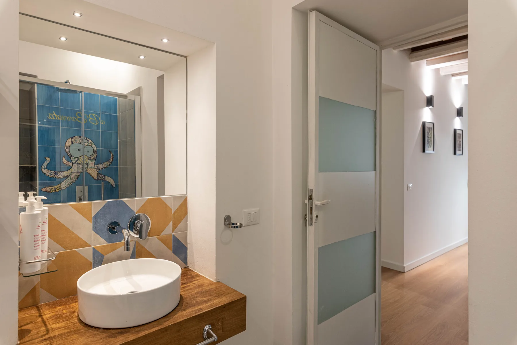 A bathroom vanity with a round basin stands beside a tiled mirror wall.