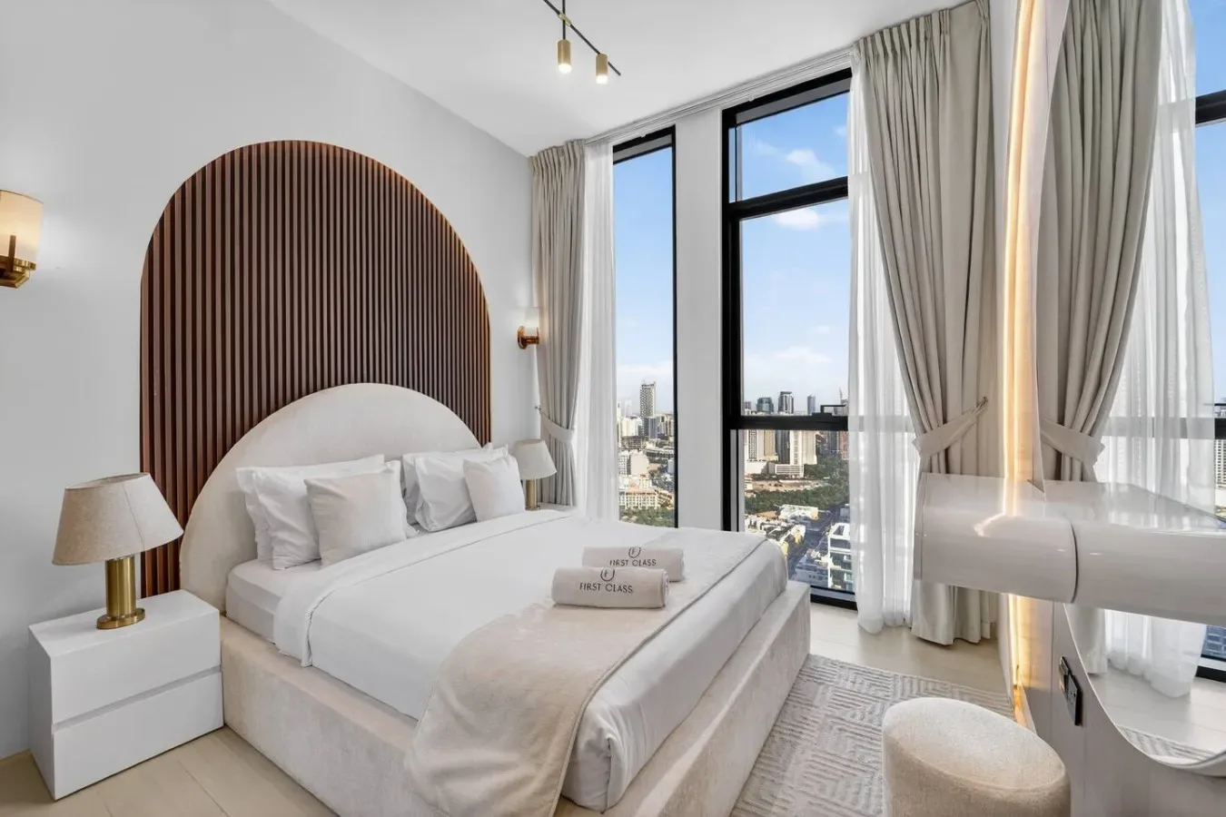 A bright bedroom with a neatly made bed and tall windows overlooking the city.