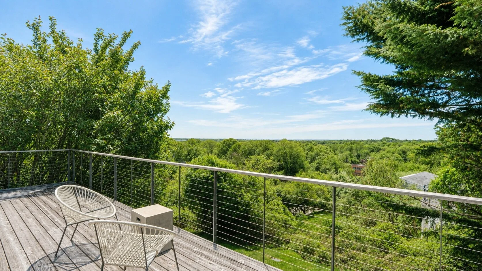 An outdoor balcony has wire railing, seating, and a view of green trees.