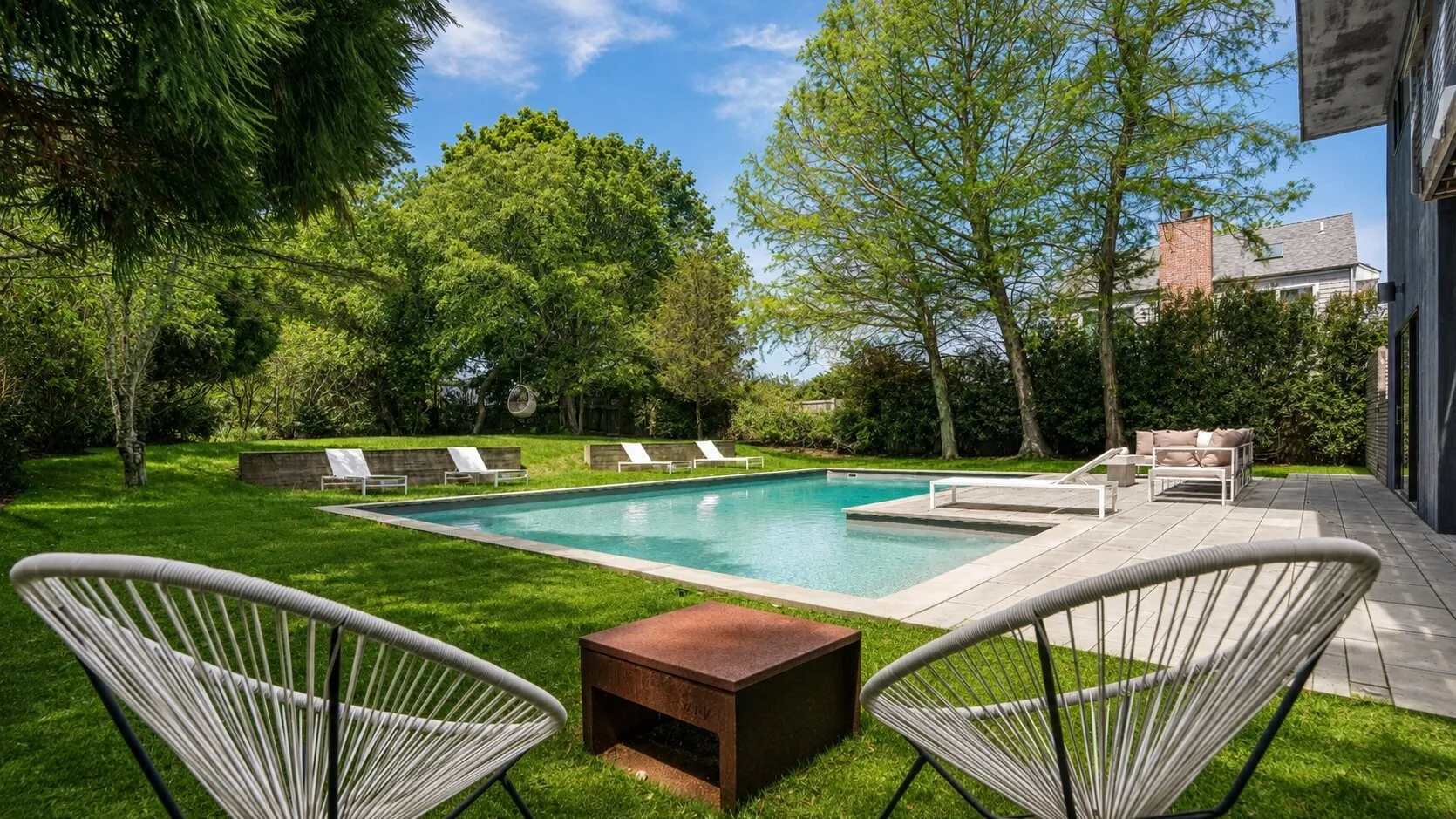 An outdoor swimming pool sits beside a lawn with seating and leafy trees.