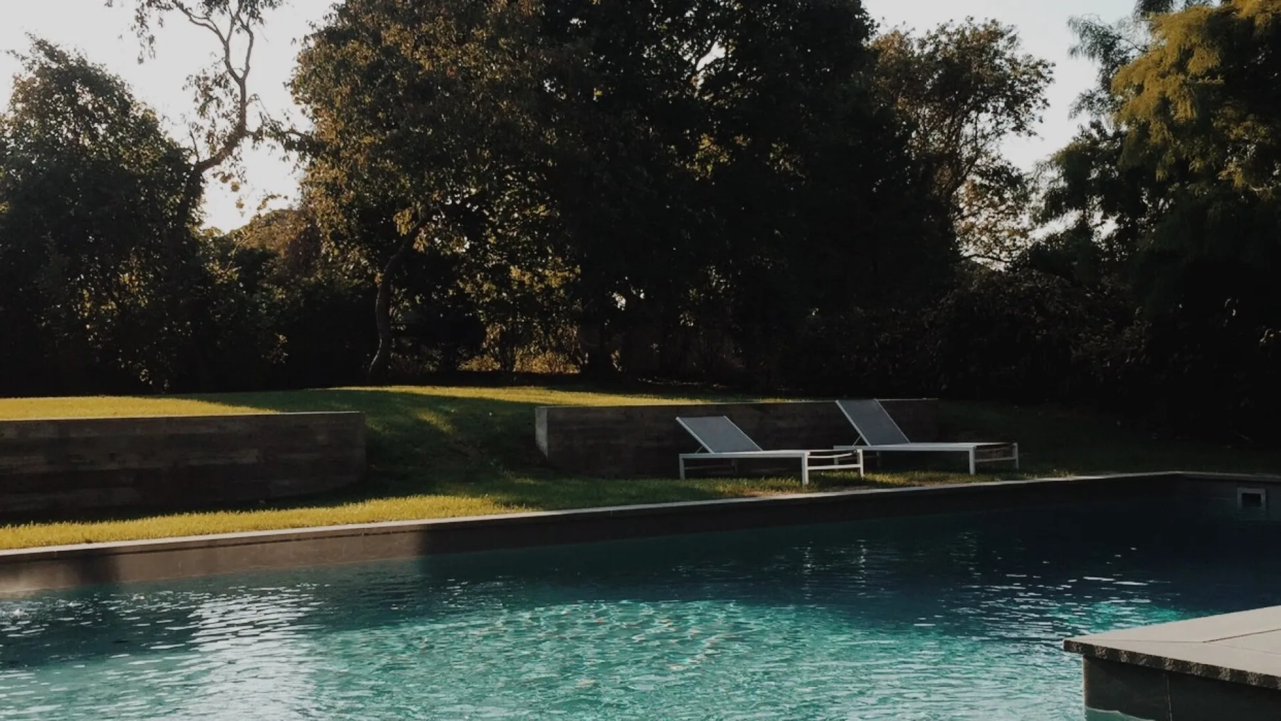 An outdoor swimming pool sits beside a grassy area and mature trees.