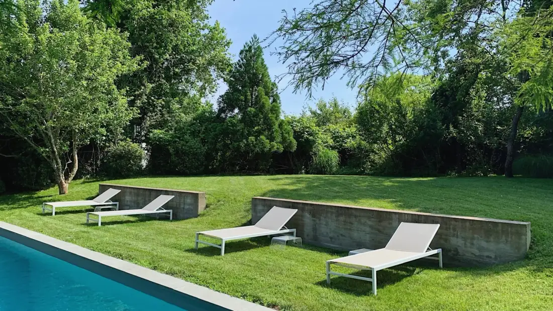 Una piscina bordea un jardín de césped con tumbonas y árboles alrededor.