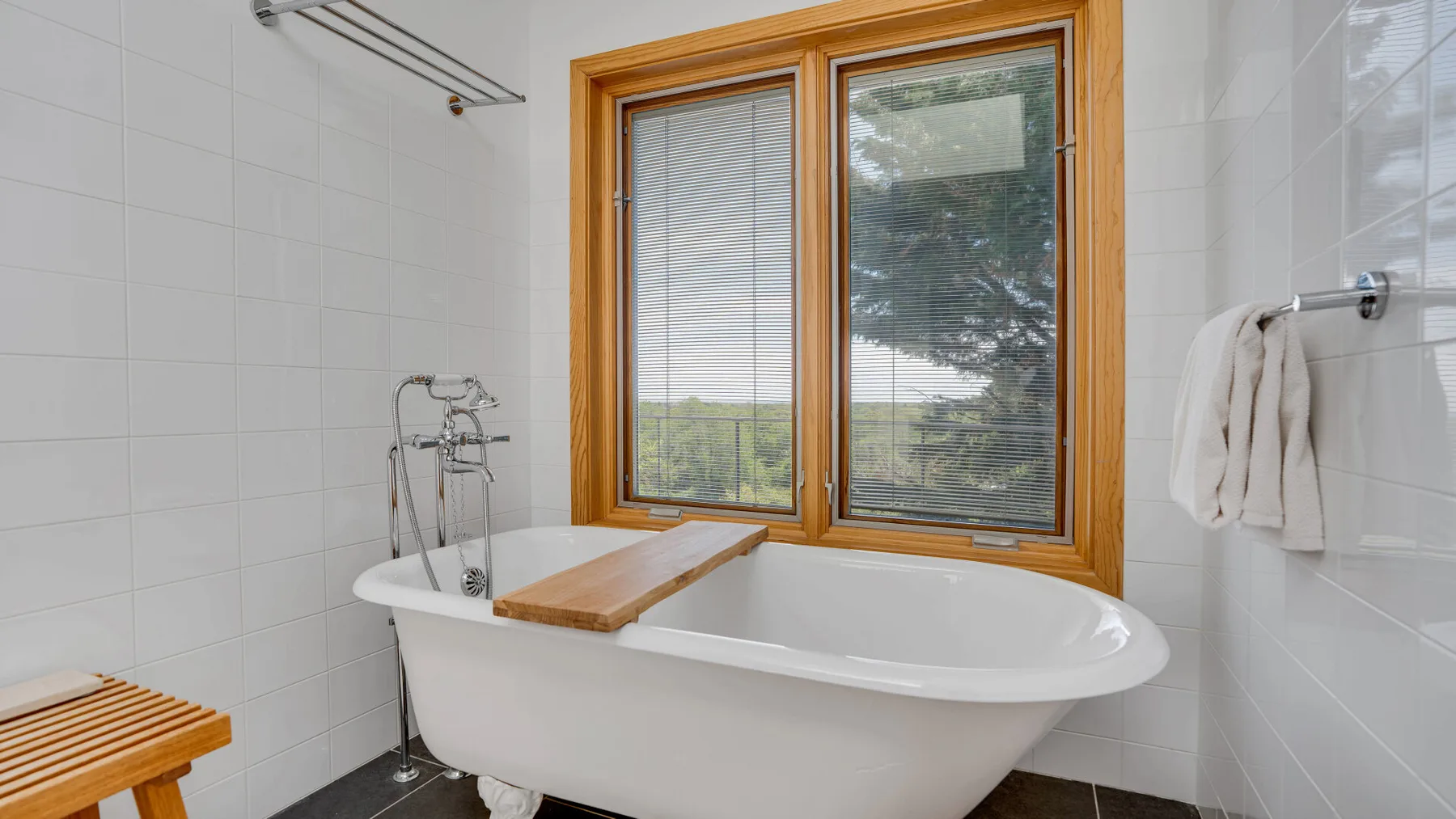A white bathtub sits beneath a wood-framed window in a tiled bathroom.