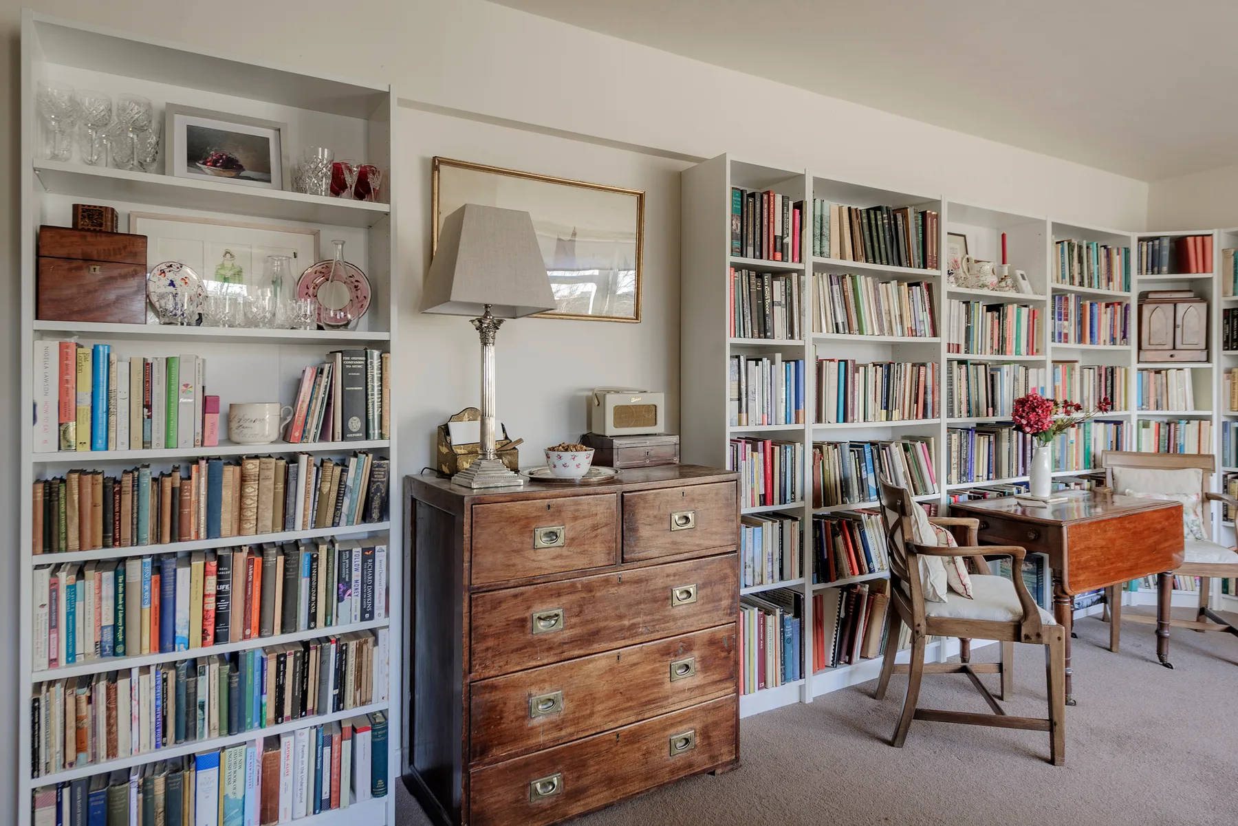 A book-lined study with a wooden desk, chairs, and a chest of drawers.