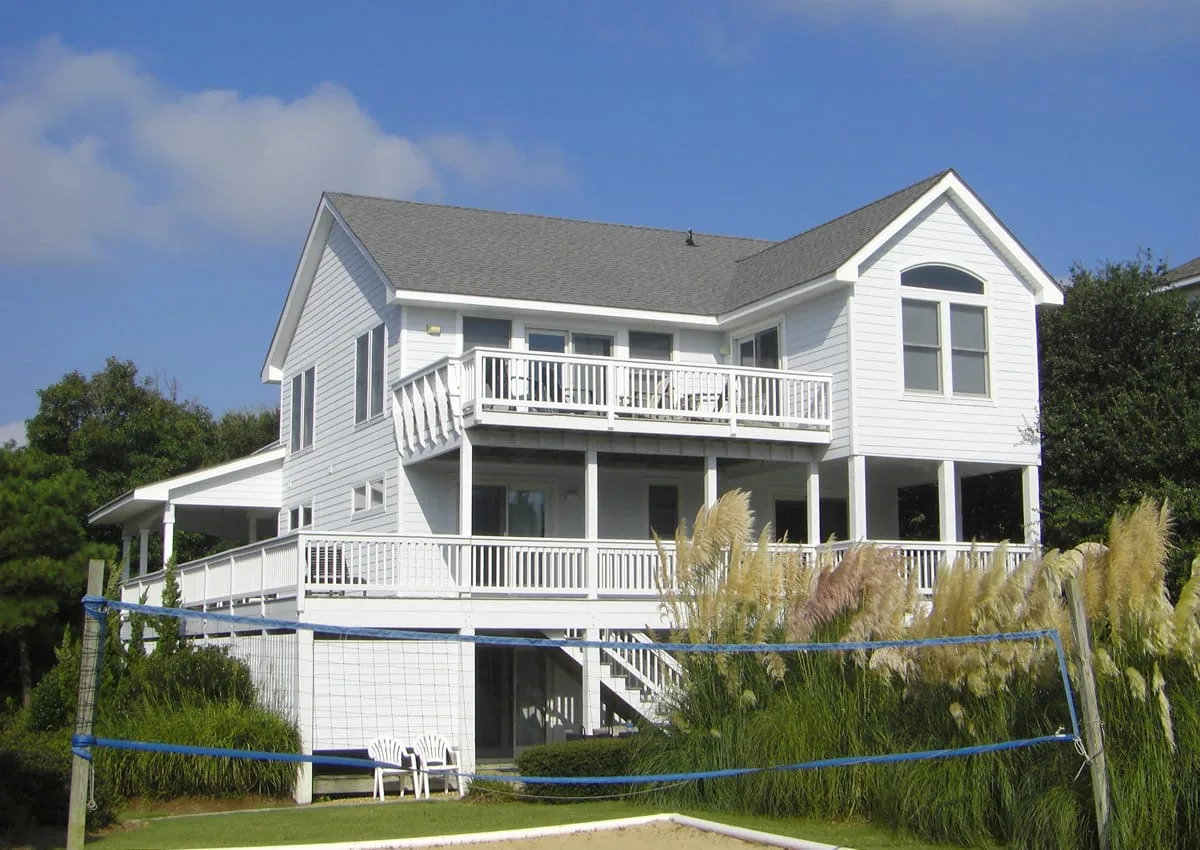 A white two-story house with raised decks stands behind a grassy yard.