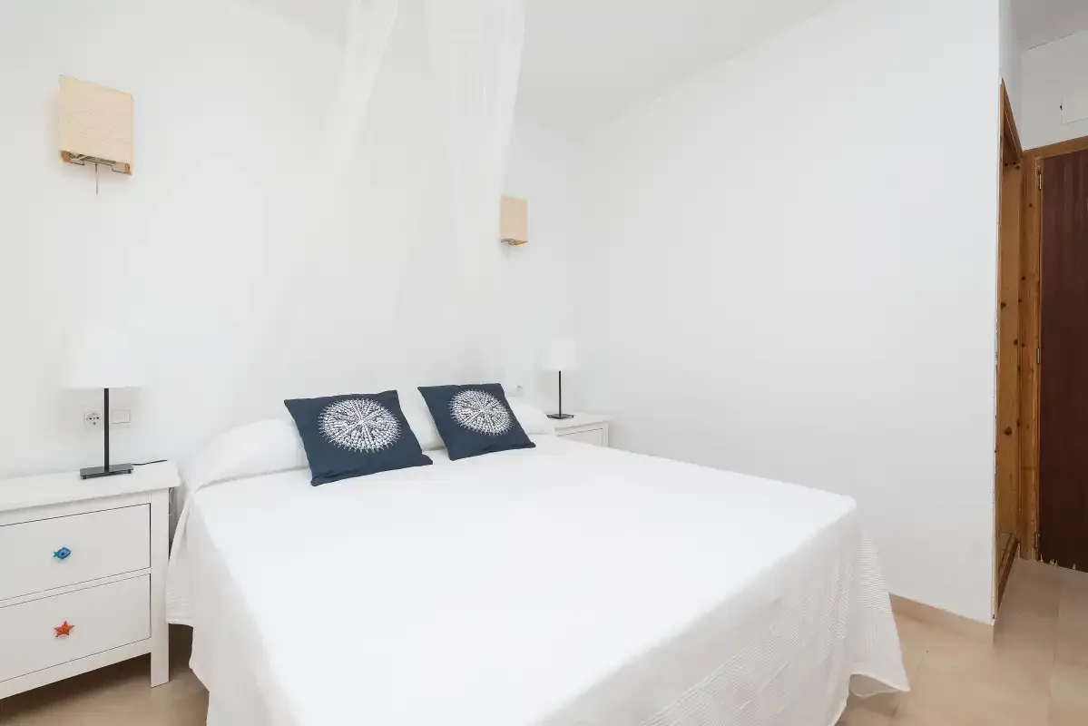 Un dormitorio luminoso con una cama blanca y cojines decorativos oscuros.