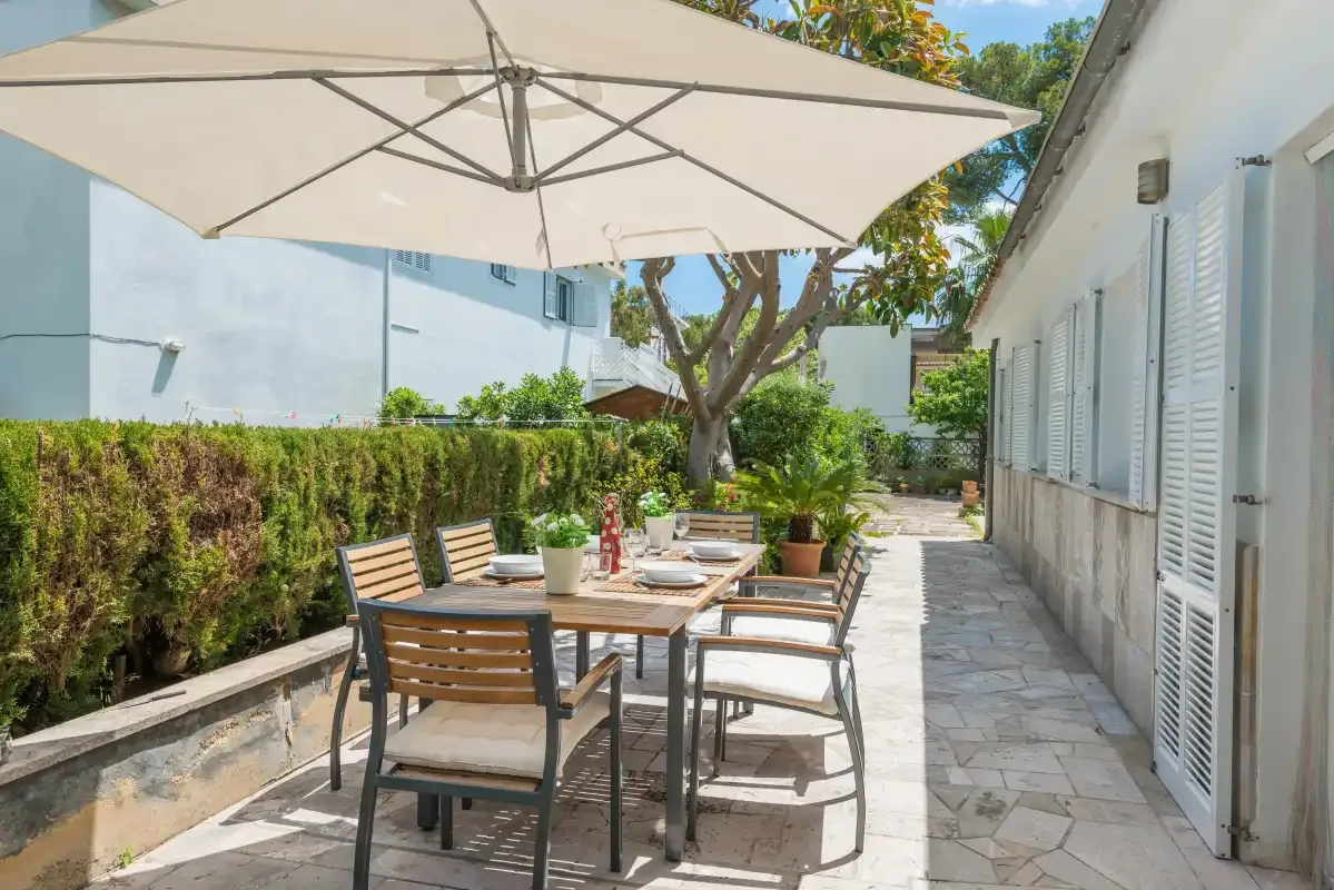 Un patio exterior sombreado con una mesa de comedor junto a un edificio blanco.