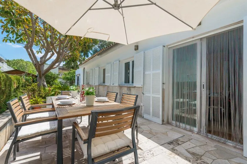 Una zona de comedor al aire libre está junto a una casa, bajo una sombrilla grande.