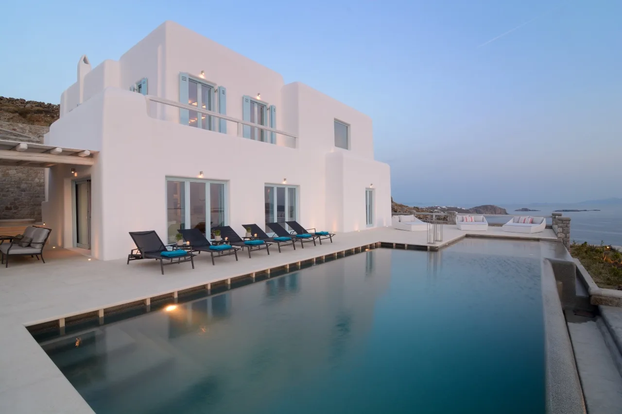 A modern white villa overlooks a swimming pool beside the sea.