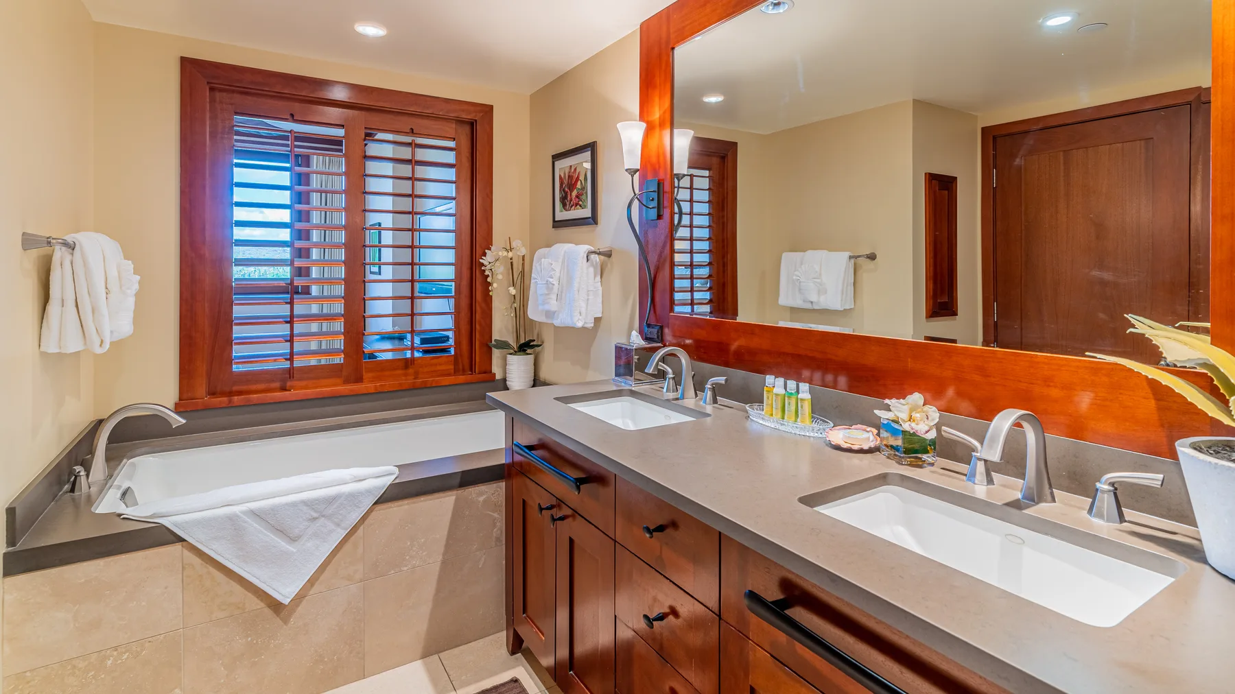 A bathroom vanity with a large mirror sits beside a bathtub beneath a shuttered window.