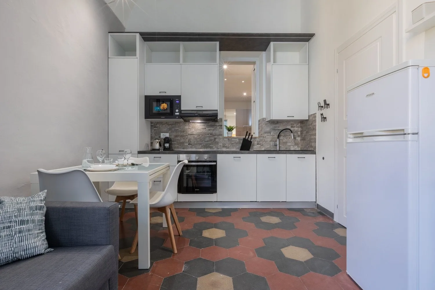 A compact kitchen with white cabinets, a dining table, and patterned floor tiles.