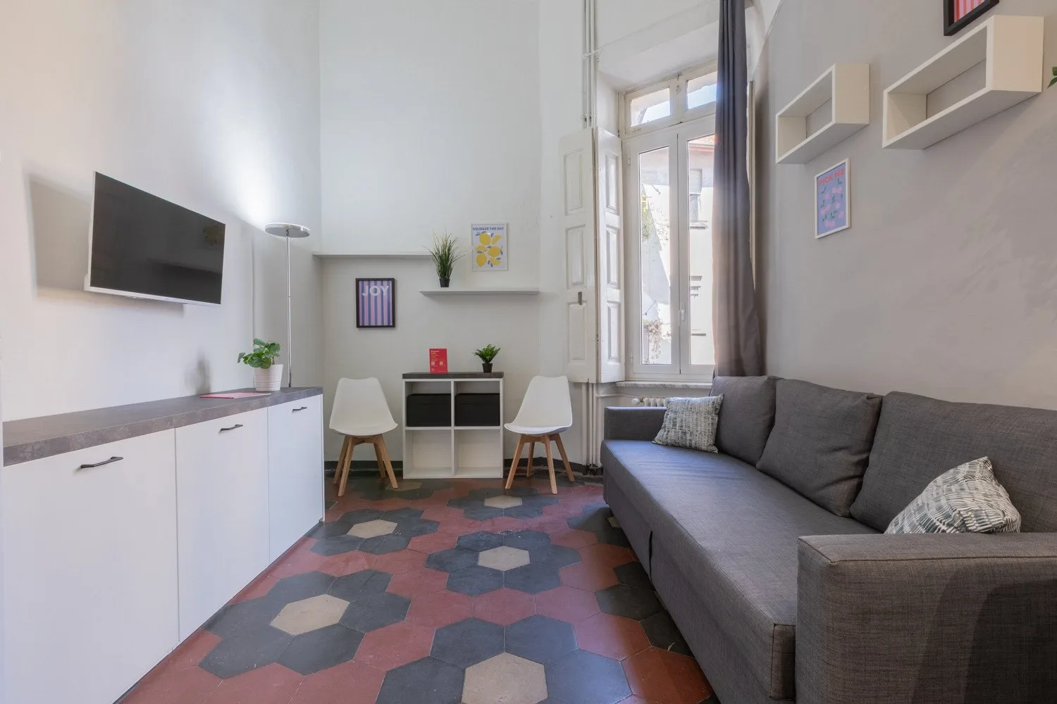 A living room with a gray sofa, wall-mounted television, and colorful patterned floor.