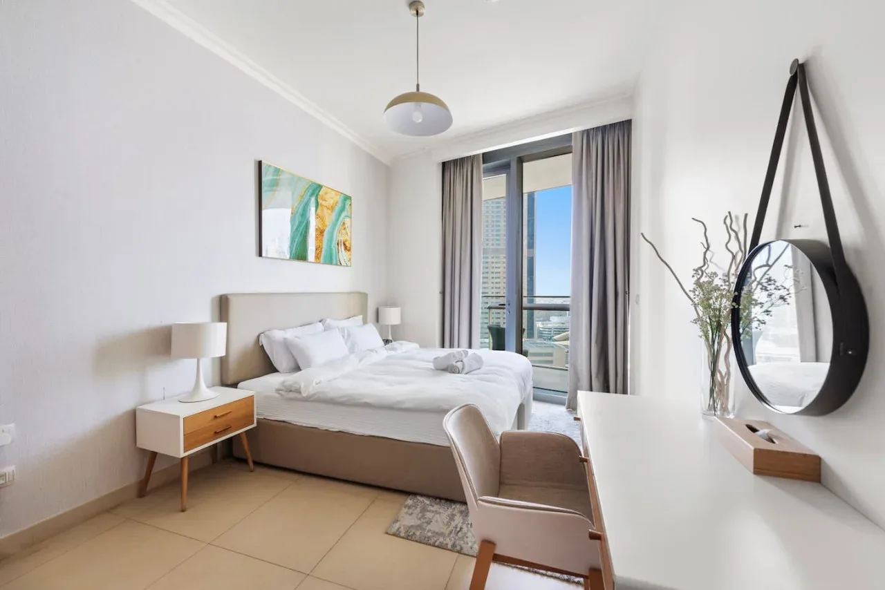 Un dormitorio luminoso con una cama bien tendida y grandes ventanales con vistas a la ciudad.