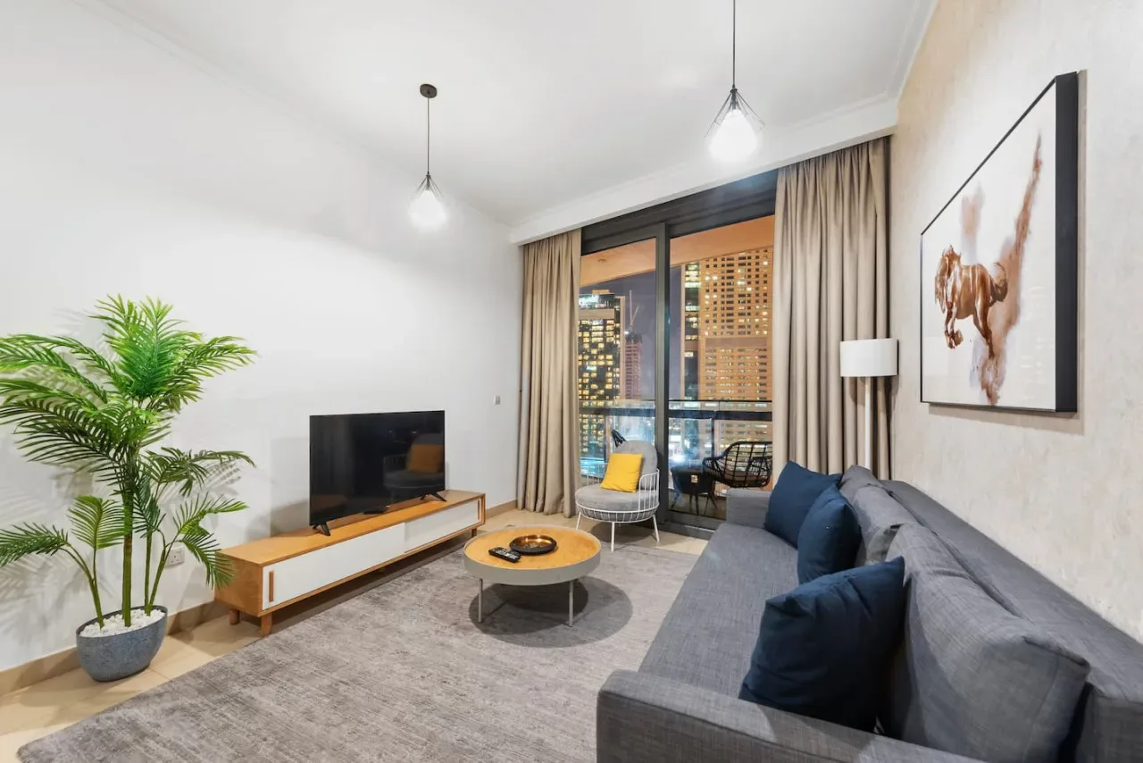 Una sala de estar con asientos, un televisor y ventanas con vista a edificios iluminados.
